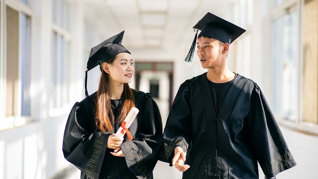 Two Chinese college students dressed in graduation gowns and caps walk through a university hallway building with diplomas in hand.