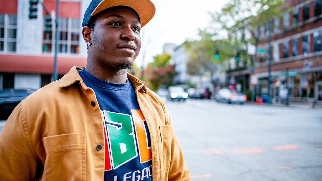 Black male HBCU college student wearing a cap and a blue sweatshirt with his school's HBCU logo walks downtown in a city on a fall afternoon.