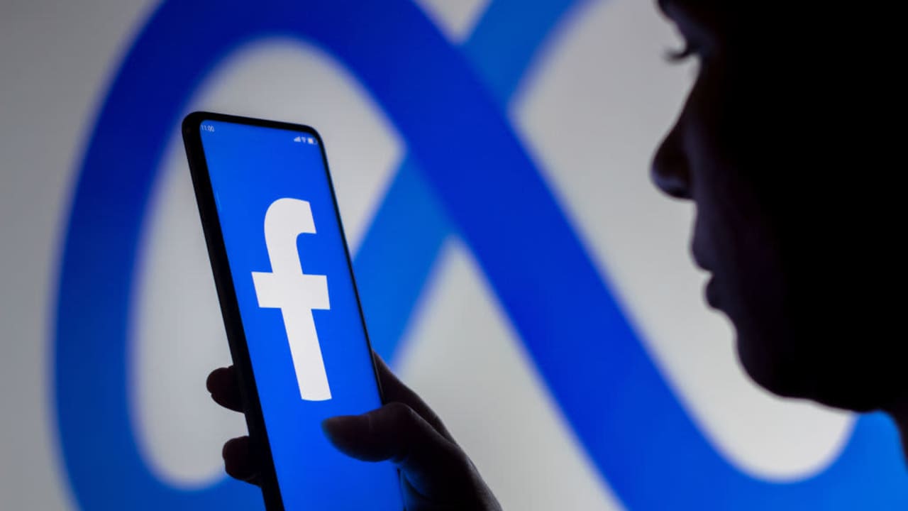 A woman standing in front of a Meta Platforms brand sign holds a smartphone with the Facebook logo displayed on the screen. The Department of Education has been tight-lipped since a report revealed that FAFSA applicant info may have been shared with Meta.
