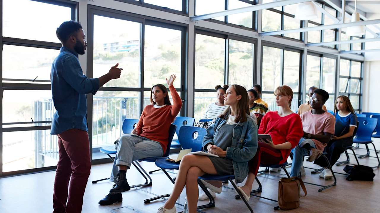 Young male professor explaining multi-ethnic students sitting on chairs by window in classroom at university