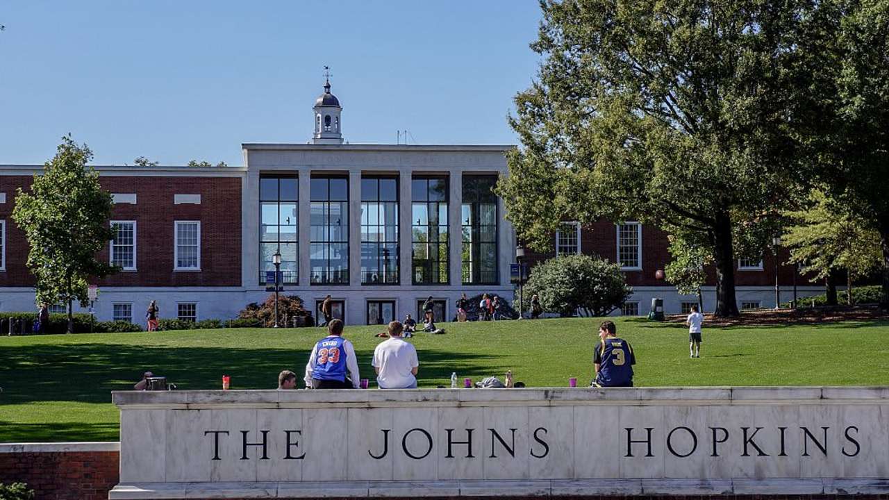 A view from Charles street of the grassy "Beach" and Milton S Eisenhower Library of the Johns Hopkins University; three male students, their backs facing the camera, sit on top of the marble university sign, other students sunbathing and playing catch on the grassy hill; the clock tower of Gilman Hall, a humanities building, peeks above the top of the library; Baltimore, Maryland, March, 2014. Courtesy Eric Chen. (Photo by JHU Sheridan Libraries/Gado/Getty Images).