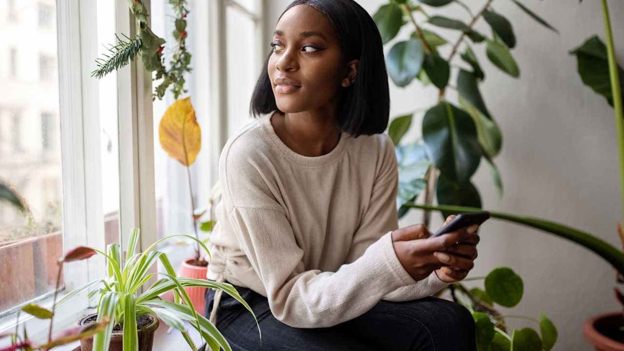 Woman sitting on the windowsill with mobile phone and looking away. Woman relaxing at home looking outside the window.