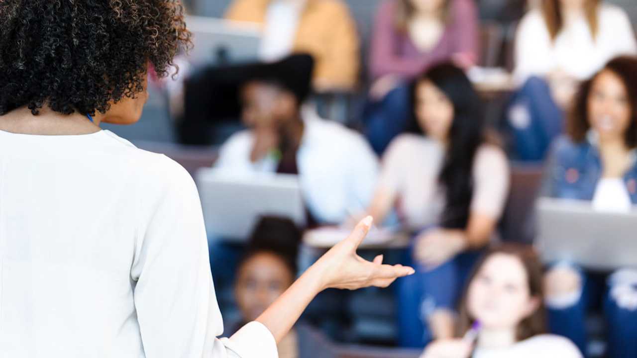 An unrecognizable mature adult female professor gestures while teaching a diverse group of college students.
