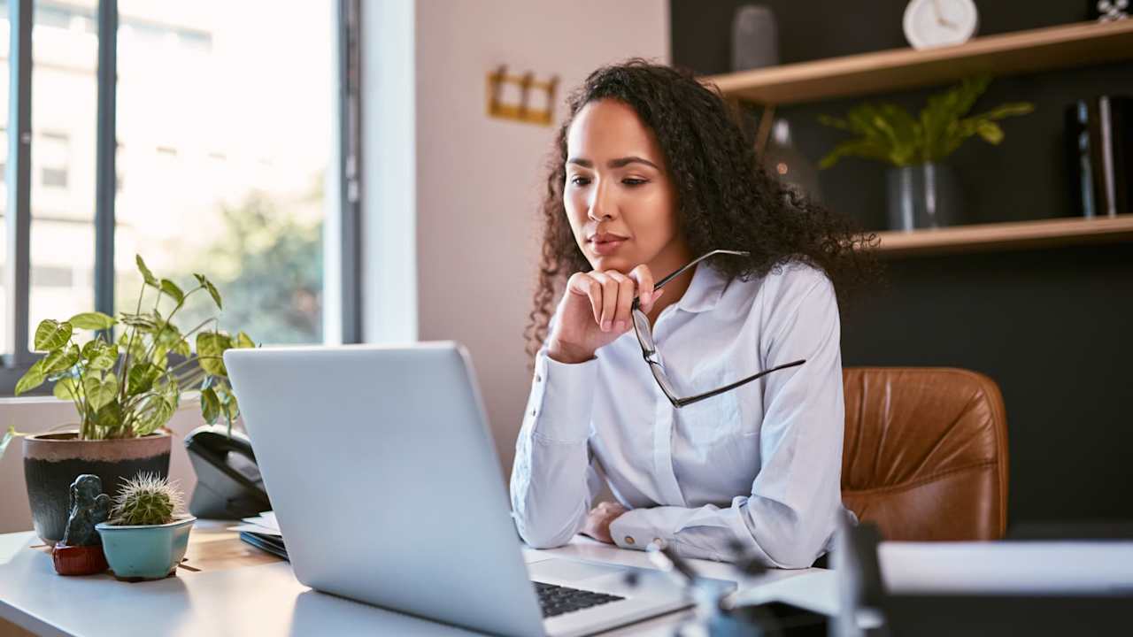Female accountant sitting at a desk in her office. She has taken her glasses off, and is reading documents on her laptop computer.