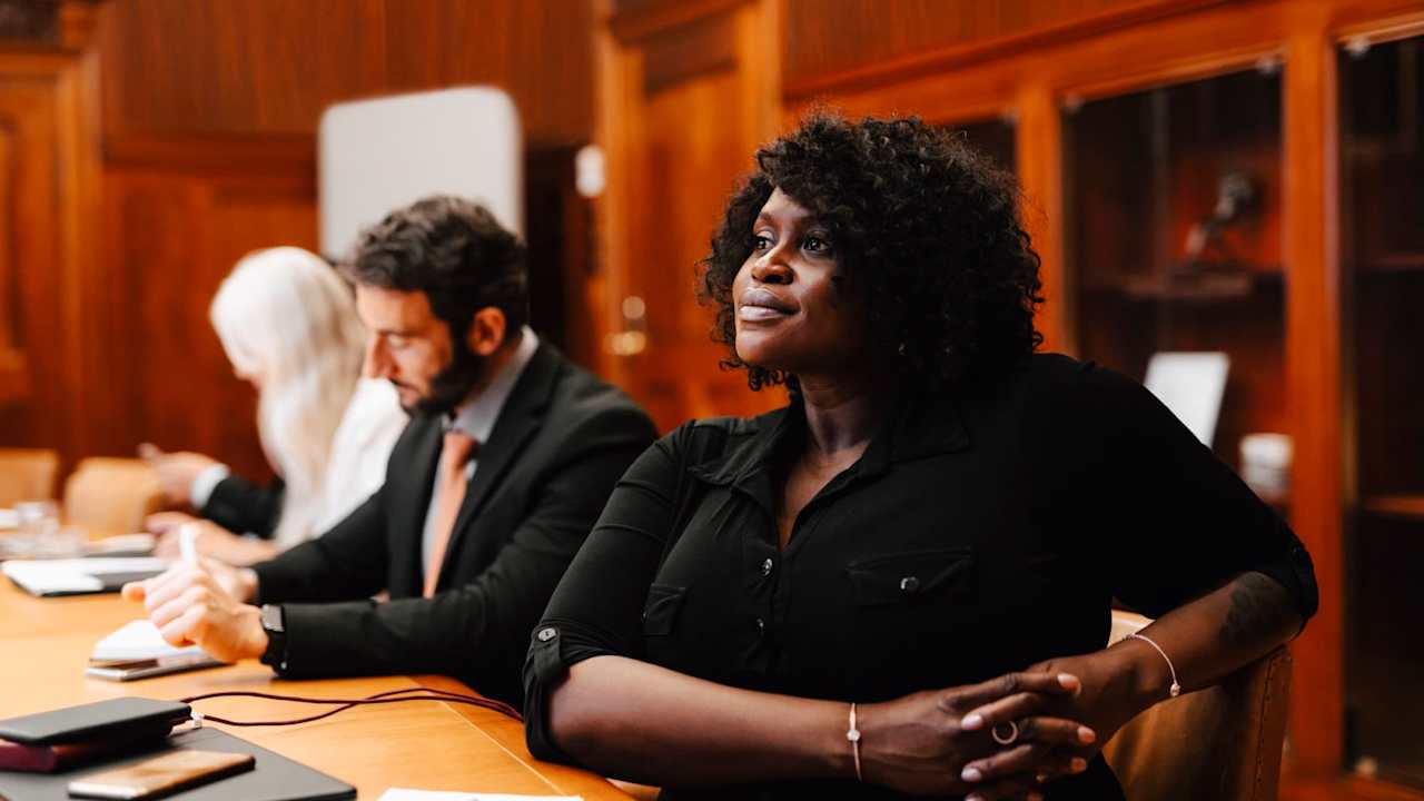 Black female lawyer sitting in a meeting room with colleagues. She is listening intently to the speakers across from her (not pictured in the photo). She is wearing a black formal suit, and has notepads resting on the table in front of her.