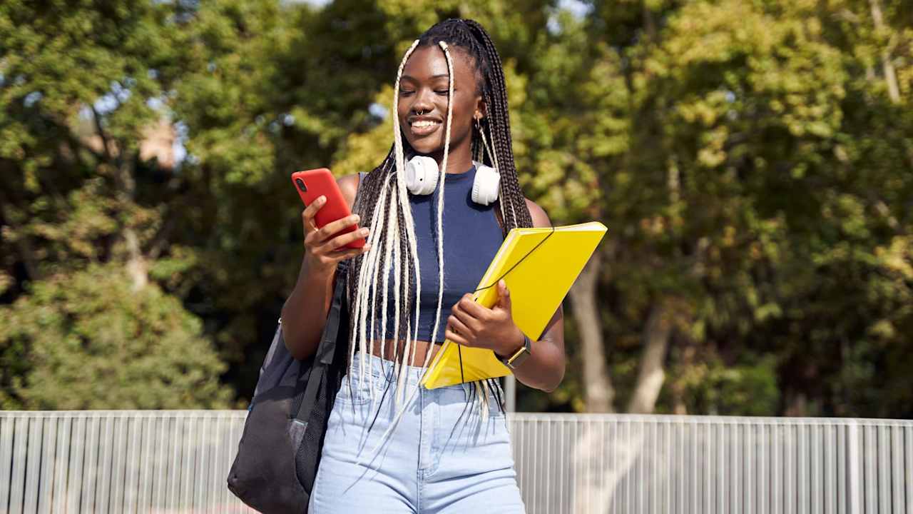 Cheerful young Black female college student standing outdoors in the city with backpack and folders in hand while using her mobile phone.