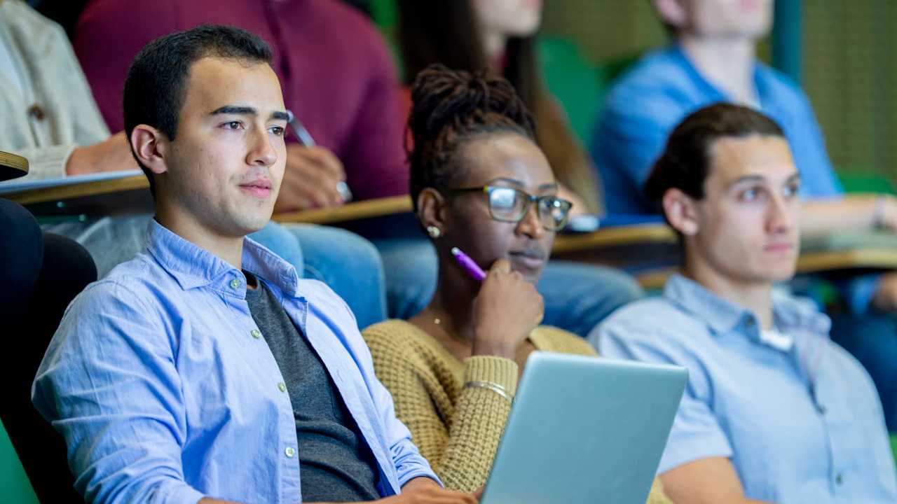 A diverse group of college students are sitting in a university classroom. They are taking notes while listening to their professor (located offscreen) during the lecture.