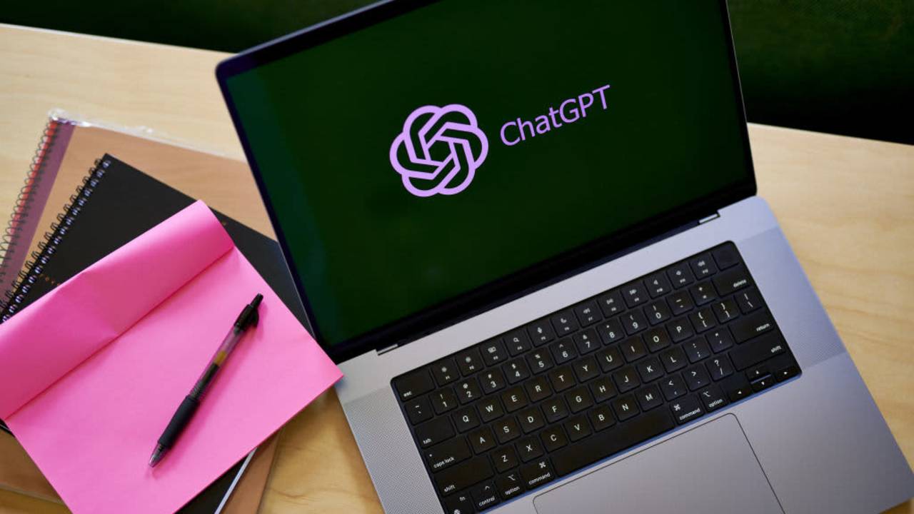 The ChatGPT app displays on a laptop computer resting on a cafe table. A notepad with a pen is resting to the left of the computer.