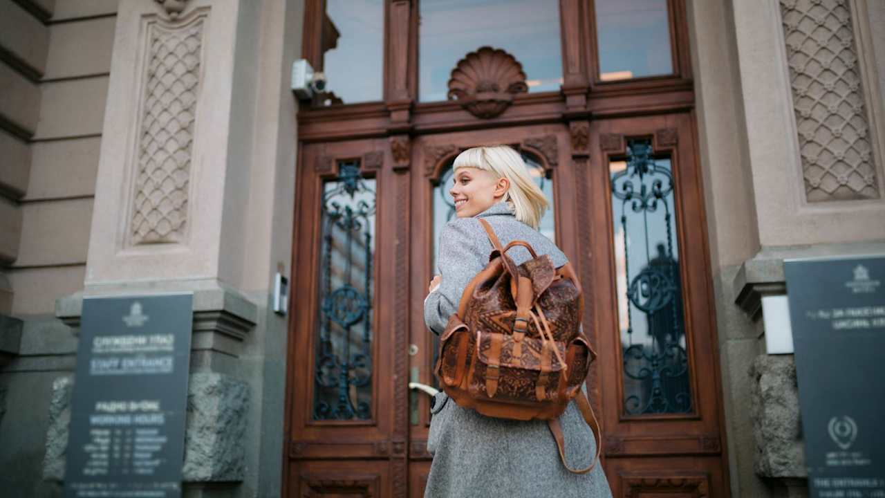Female university student with blond hair walks towards a building entrance on campus with her backpack and smiles.
