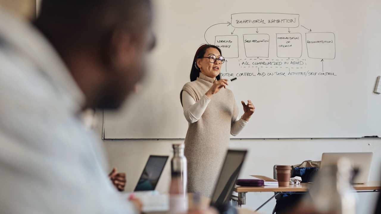 Asian American female college professor gestures with a marker while explaining a diagram on a whiteboard behind her to students in the classroom.