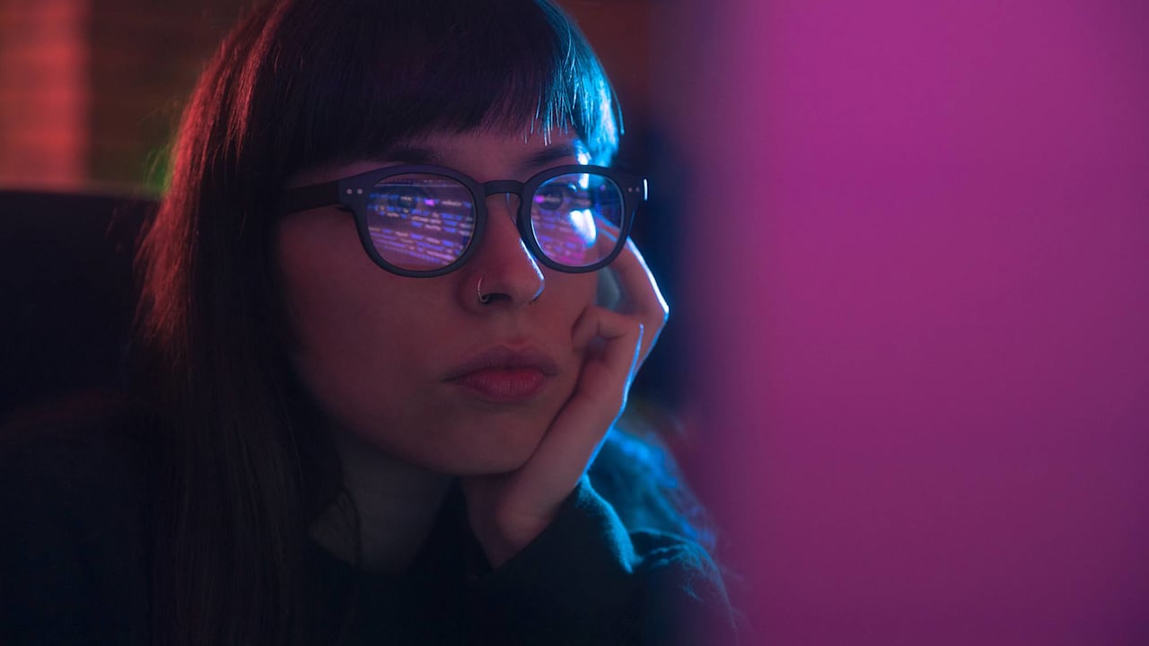 Caucasian female programmer wearing glasses is coding on her desktop computer. She is resting her head on her hand.