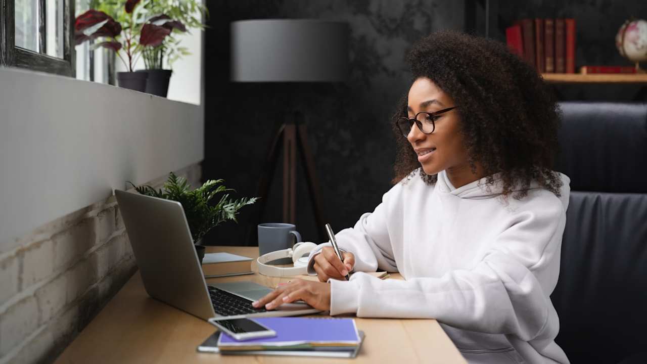 Black high school student sitting in an office at home. She is typing on her laptop computer with her left hand, while taking notes on a notepad in her right hand.