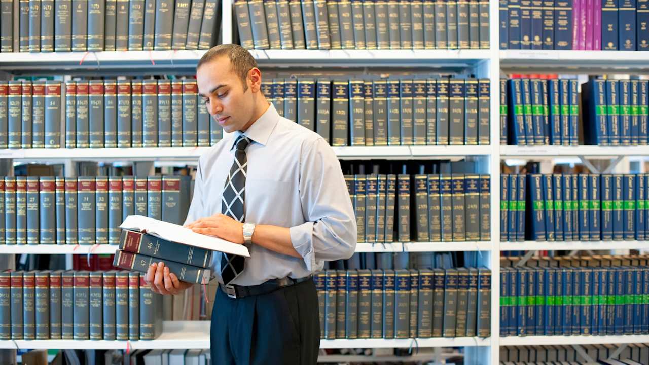 South Asian male law student reading a legal textbook in the library stacks on campus.