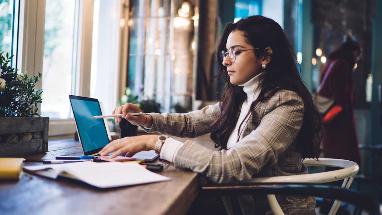 Female law student wearing glasses sitting at a table in a cafe. She is writing notes on a digital tablet. She is studying from several open textbooks.
