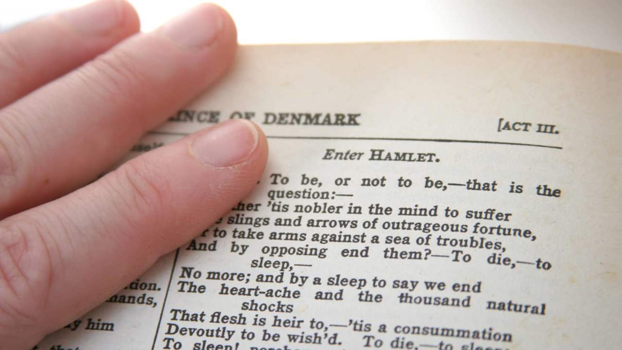 hand resting on a page of a book next to the lines: to be or not to be, close up