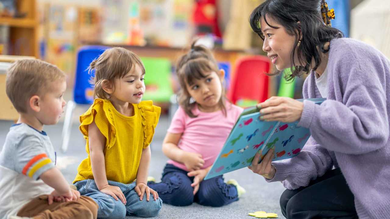A preschool teacher sits on the floor of her classroom with a small group of students as she reads them a book.