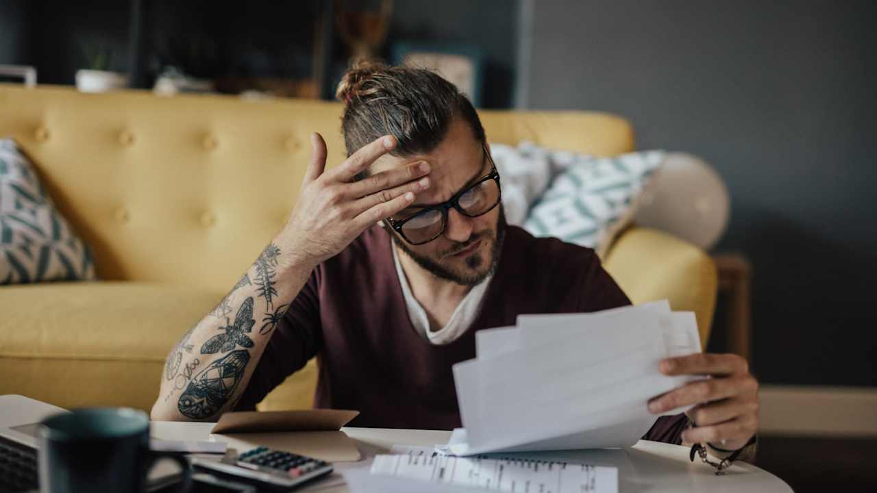 Caucasian male college student sitting at his living room coffee table. He has a worried look on his face while he's holding several paper bills in his hand.