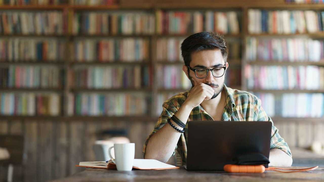 Caucasian male student reviewing his homework assignment on a laptop. He is sitting at a table in a university library, with an open textbook and a coffee cup next to him.