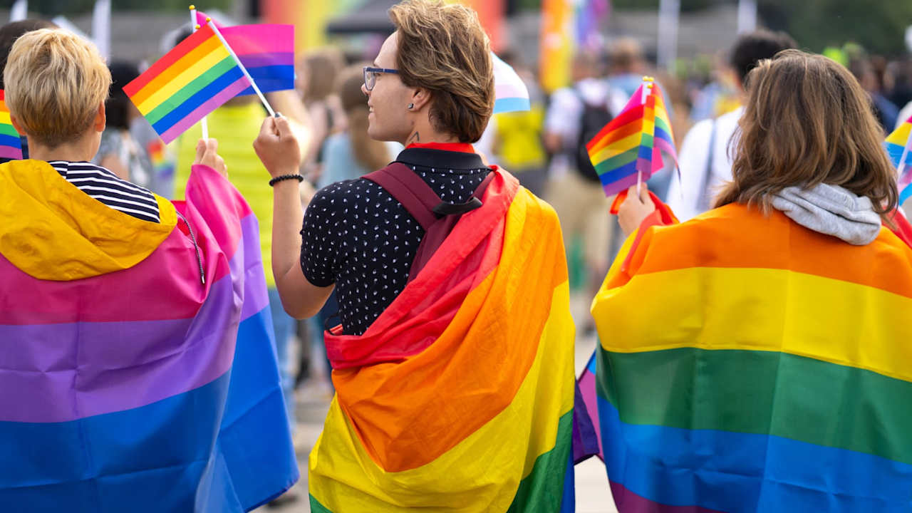 A group of three college students wrapped in bisexual and gay pride flags are waving small flags while watching a gay pride parade during Pride Month.