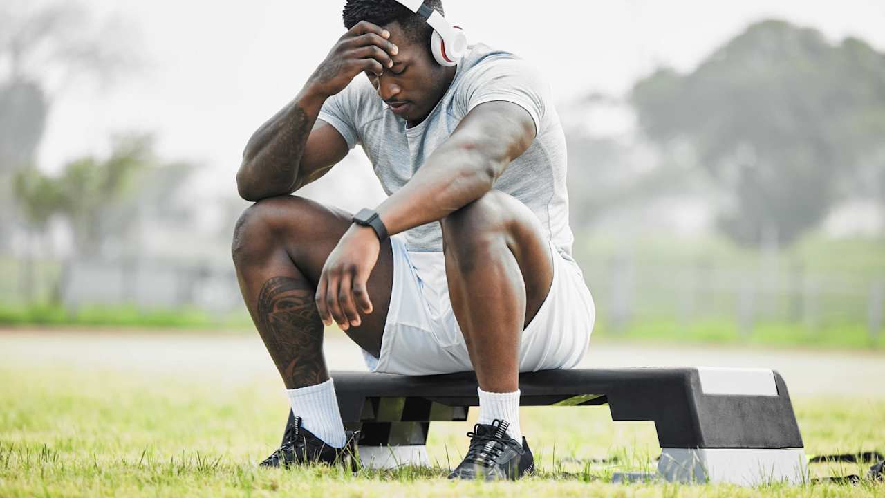 Black male college football player sitting on the field. He is rubbing the temples on his forehead, and has an exhausted look on his face.