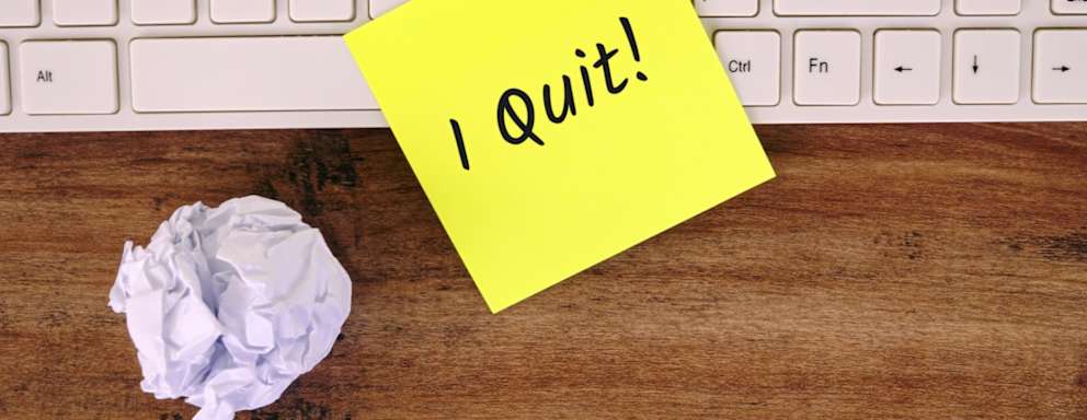 Post-it note with the words, "I Quite!" written on it is placed on top of a keyboard on a worker's office computer.