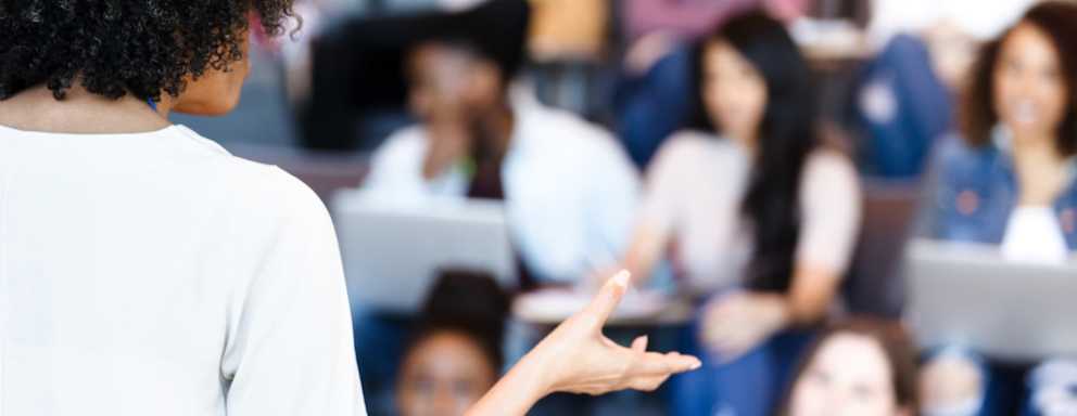 An unrecognizable mature adult female professor gestures while teaching a diverse group of college students.