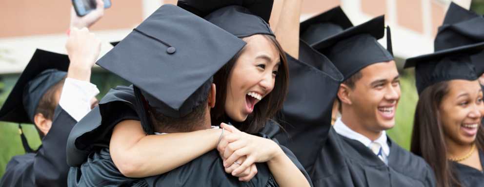 A group of college students dressed in caps and gowns congratulate and hug each other on graduation day.