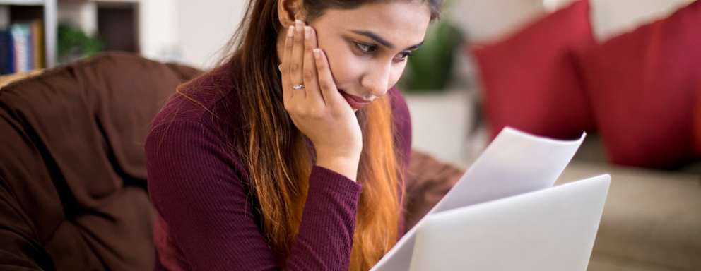 Female college graduate sitting on a couch at home going through her student loan bills with a worried look on her face.
