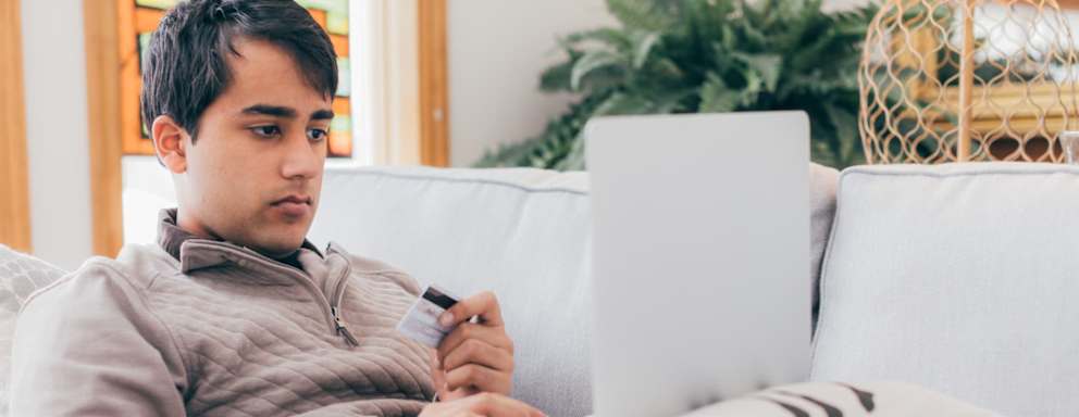 College student lying on a couch in his living room paying his student loan bill online with his credit card.