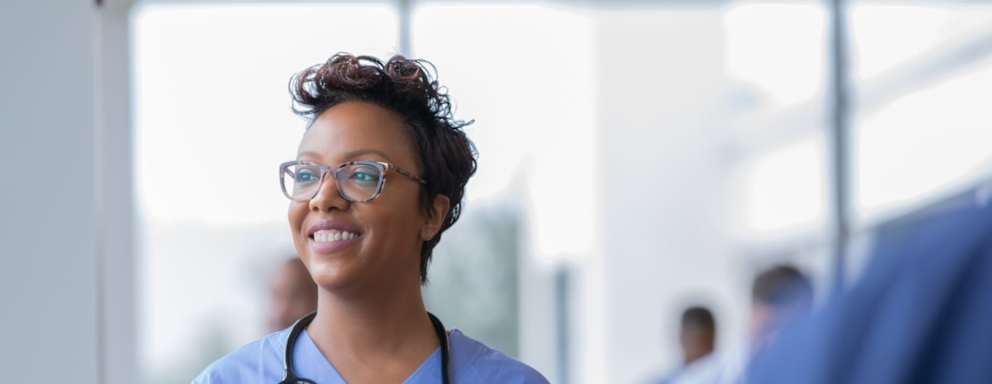 Female nurse smiles while staring out window in hospital hallway and holding digital tablet with electronic patient file