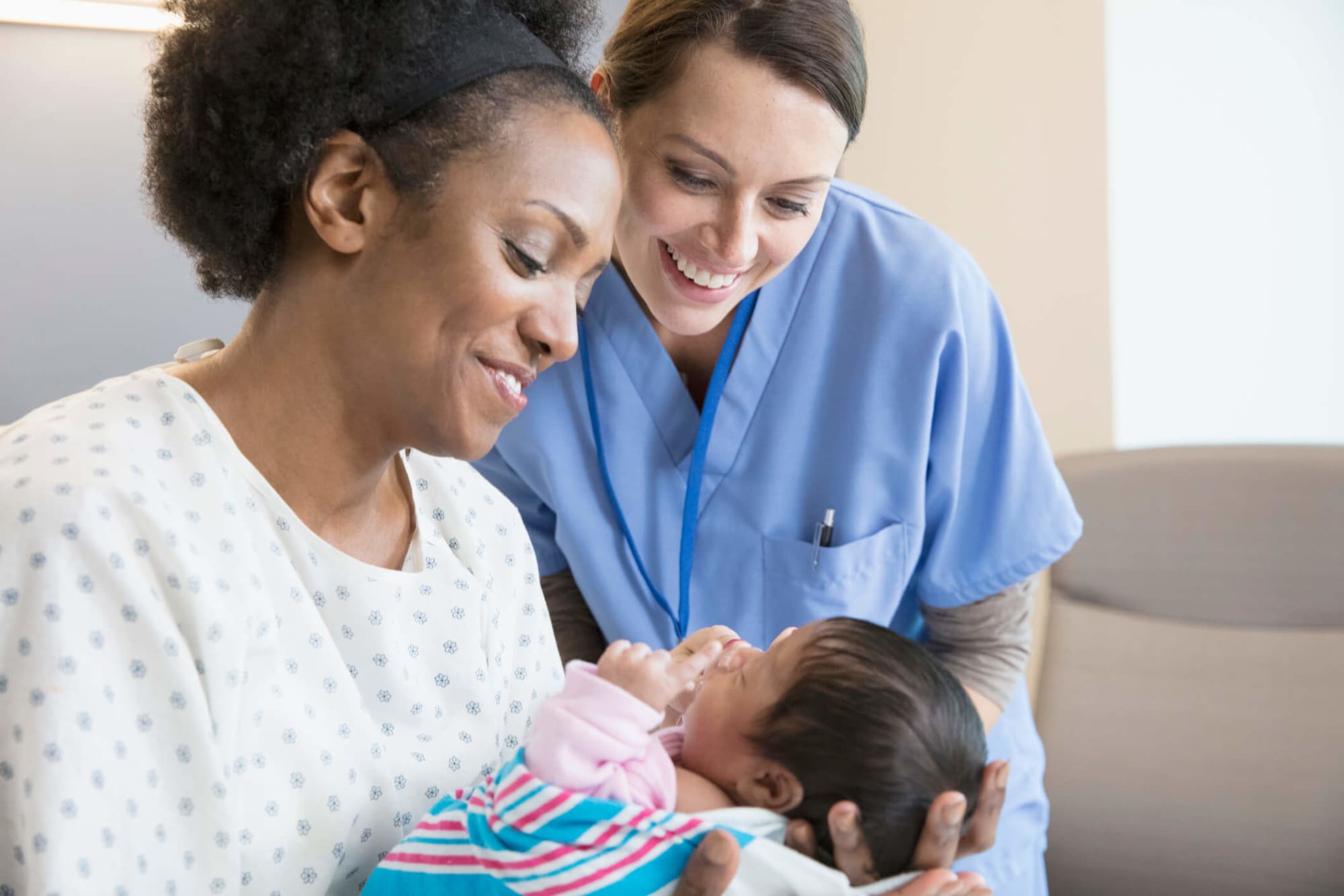 A mid-adult Caucasian nurse midwife is checking in with an African-American mother and her newborn child. The mother is lying in a hospital bed, swaddling her baby in her arms and smiling. The midwife is wearing light blue scrubs. She is standing over the mother and baby and smiling while she does her health check-in with them.