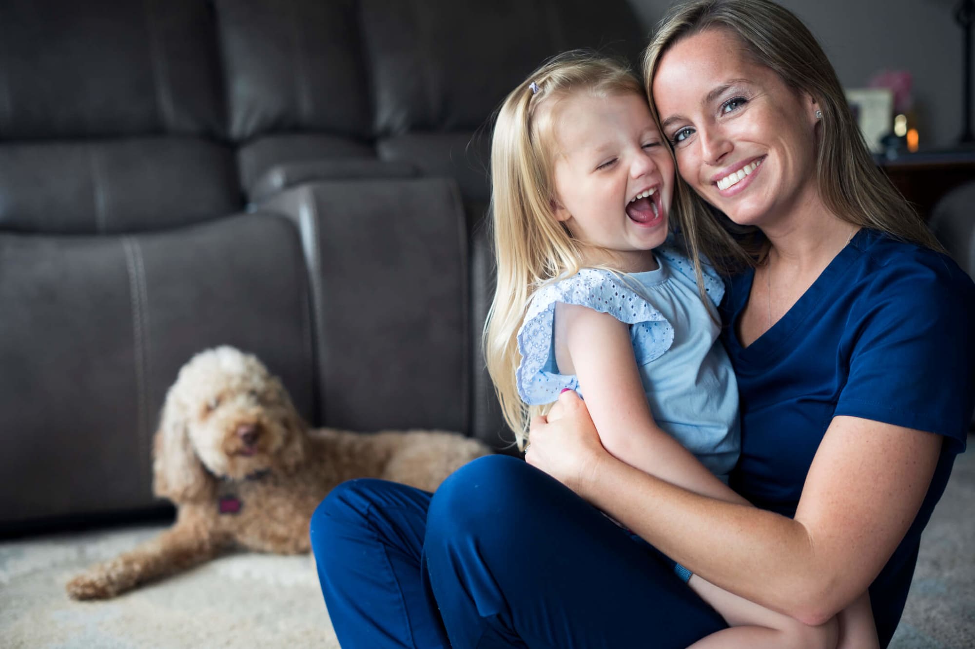 A mid-adult Caucasian nurse wearing dark blue scrubs is enjoying some bonding time with her little daughter before work. She is sitting down ion the floor of their living room, holding her daughter in her arms and smiling. The young daughter is wearing a light blue top and laughing with her eyes closed