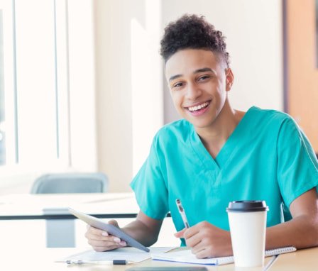 Black male nursing student sitting at a classroom desk writing notes while preparing for class. He is wearing scrubs and referencing materials from a digital tablet.