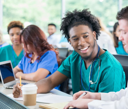 A young African American nursing school student is discussing class material with his instructor. The student is wearing teal green scrubs, and is taking notes and smiling as he listens to his instructor explain. The instructor is kneeling next to him and showing him something on a notepad. Other students are studying in the background.