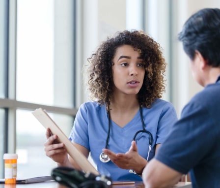 A Black female nurse case manager is discussing a care plan with a male patient. They are sitting at a desk in a hospital office. The nurse is showing the patient an outline of the plan on a clipboard.