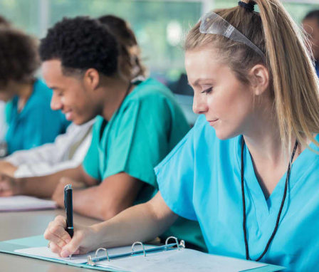 A young Caucasian female nursing student is taking notes during her professor's lecture. She is sitting down at a desk and wearing light blue scrubs. Her classmates are pictured behind her taking notes.
