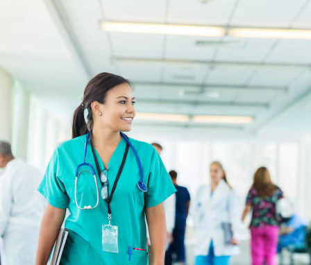 Confident young Hispanic nurse looks to the side while walking in a hospital corridor. She is wearing scrubs, stethoscope, glasses and id badge. She is carrying medical charts. Medical professionals are walking in the background.