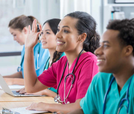 Smiling young female African American student raises her hand to ask a question in class. She is sitting in the middle of three other students at a string of desks. Two other students are looking forward and smiling and another is writing at her desk.