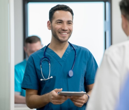 Young adult Hispanic man is smiling while talking to coworker in hospital emergency room. Nurse or doctor is holding a digital tablet.