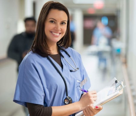 Female Caucasian nurse standing in a hospital hallway writing note on her clipboard. She is turning her head toward the camera and smiling.