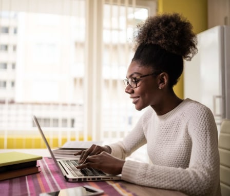 A college-aged African-American woman is studying for class at home. She is wearing a white wool sweater and glasses, and has her curly dark brown hair pulled up into a high ponytail. She is sitting at her kitchen dining table using her laptop to study. There are also a few books and her cell phone nearby.