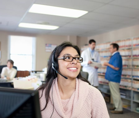 A young female medical receptionist sitting at her desk while talking with a client. Two doctors are standing while chatting in the background.