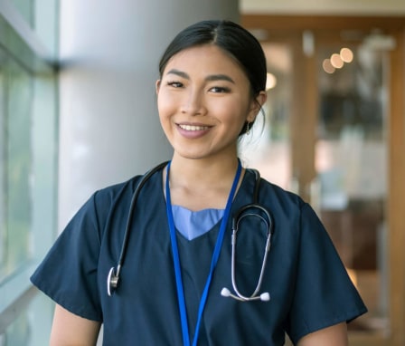 An Asian-American female nurse wearing scrubs and a stethoscope poses next to a window in a hospital corridor and smiles at the camera.