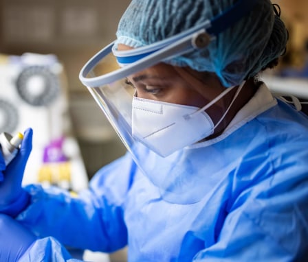 Black female epidemiologist working in a laboratory. She is wearing a personal protective suit, mask, and face shield while gathering pathogen samples.