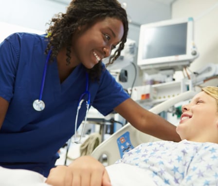 A mid-adult Black female nurse is checking in with one of her patients in the emergency room. She is wearing dark blue scrubs and has a stethoscope around her neck. She is leaning over the patient and smiling while checking his health vitals. The patient is a young Caucasian boy wearing a standard hospital robe while resting in bed. He is smiling back at the nurse, and is pictured with hospital equipment and a window behind him.