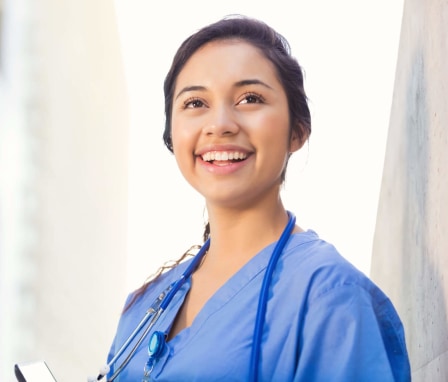 A young adult Hispanic woman is smiling while standing outside on a college campus. She is a nursing student. She is smiling and wearing blue hospital scrubs. She is also wearing a stethoscope and school ID.