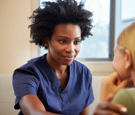 A mid-adult Black nurse comforts a young distressed female assault victim in a clinical office. The nurse is wearing dark blue scrubs, and is gently placing her right hand on the victim's left shoulder to offer her support. The victim is pictured in the foreground leaning her head on her left hand. She is a young white woman with blond hair wearing a light green shirt.