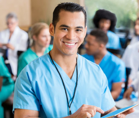 Mid-adult Hispanic nurse holds a digital tablet during a medical staff conference. He has short black hair and is wearing light blue scrubs. He is smiling at the camera. His colleagues are conversing with themselves in the background.