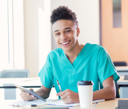 Black male nursing student sitting at a classroom desk writing notes while preparing for class. He is wearing scrubs and referencing materials from a digital tablet.