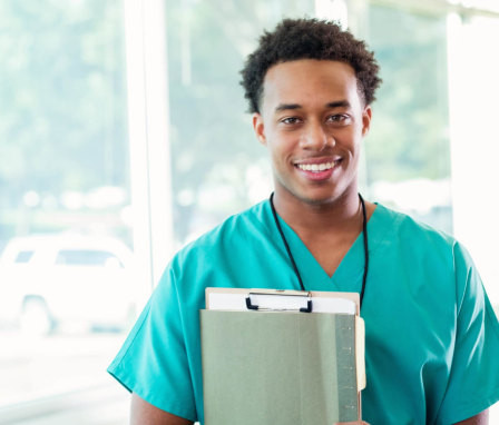 A young African-American nursing student stands outside a school hallway before his class begins. He is wearing teal green scrubs, and carries a notebook and a few file folders in his arms. He is looking at the camera and smiling.